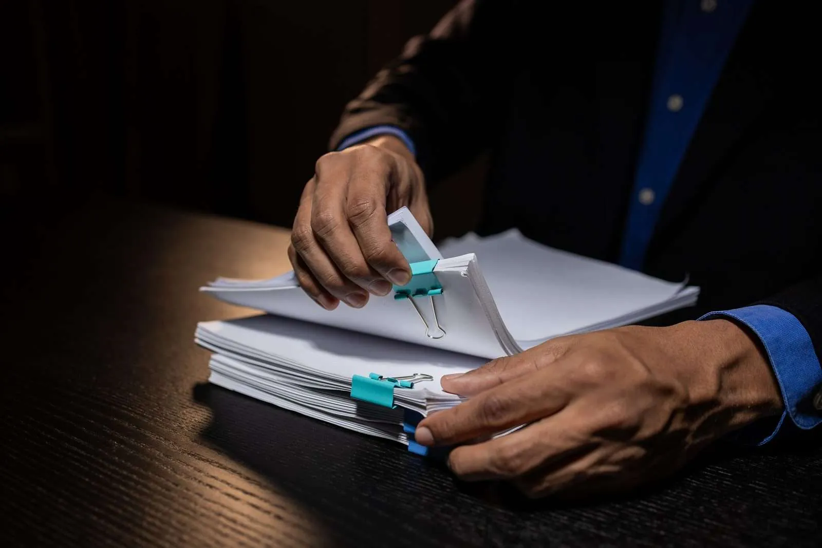 Hands sorting a thick case file held with binder clips under a desk lamp